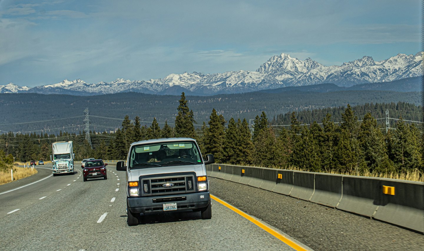 Silver Ford van drives on a highway near pine forests and mountains.